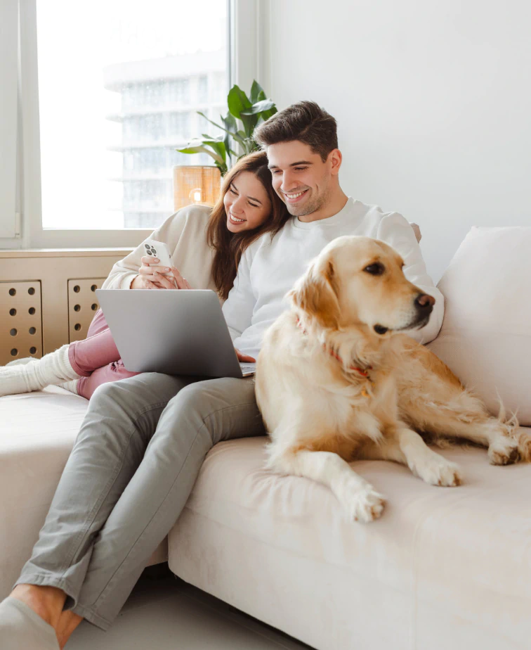 Desktop_About_Us_Insurance_Policy A couple sits on a couch, smiling at a laptop screen, with a golden retriever lying beside them. A large window and a potted plant are in the background.