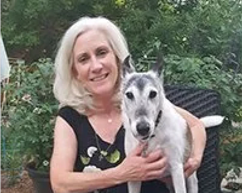 A woman with shoulder-length gray hair smiles while sitting outdoors, holding a white and gray dog on her lap. Green foliage is visible in the background.
