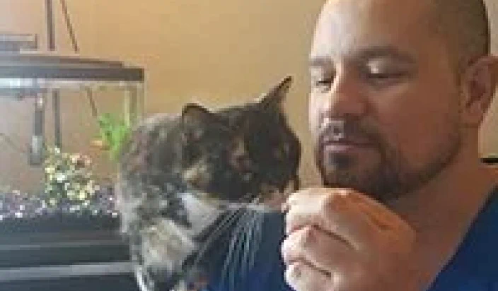 A man with a beard holds out his hand to feed a tortoiseshell cat, which is perched near a fish tank in the background.