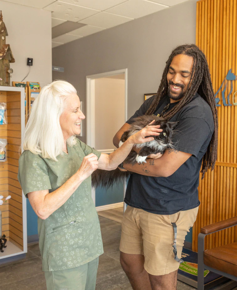 Desktop_About_Us_Insurance_Policy A smiling veterinarian in green scrubs greets a man holding a black and white cat at a clinic. The man is smiling as the vet gently pets the cats head. Shelves and wood paneling are visible in the background.