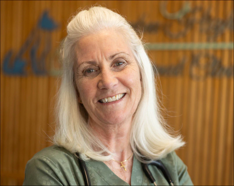 Smiling older woman with long gray hair and a stethoscope around her neck, wearing a green top, standing in front of a blurred wooden background with indistinct blue lettering.