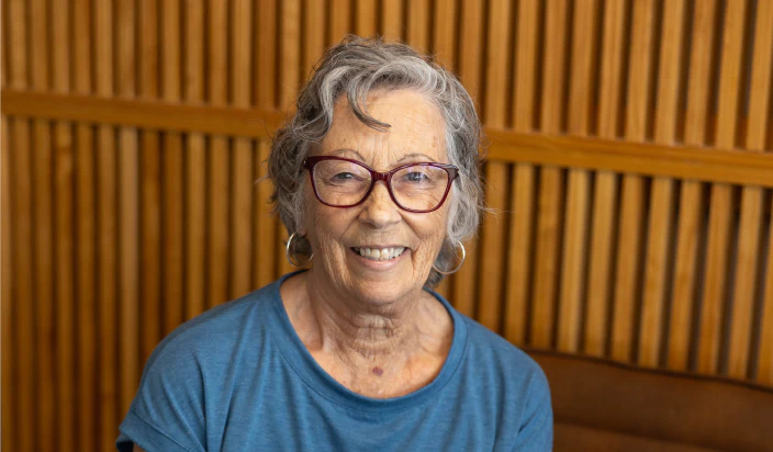 An older woman with gray hair and glasses smiles at the camera. She is wearing a blue shirt and sits in front of a wooden slatted wall.