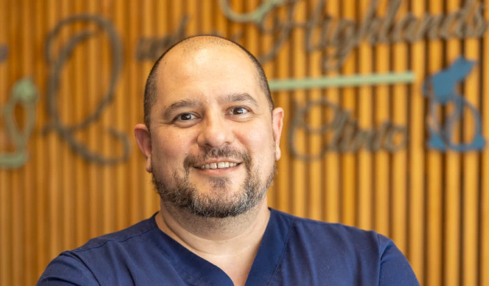 A smiling man with a beard and shaved head wearing navy blue scrubs stands in front of a wooden wall with blurred writing and symbols in the background.